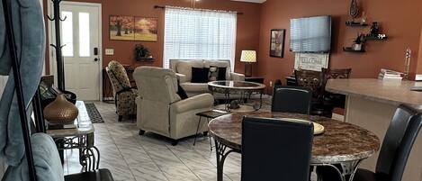 Ein Wohnzimmer mit einer Couch, einem Esstisch mit Stühlen, einem Fernseher und einer Wanduhr.