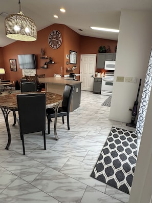 A modern kitchen with a clock, a dining area with a round table and chairs, and a patterned rug leading to a door.