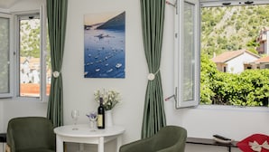 A room with a table, two chairs, a painting of a coastal scene, and a view of houses and greenery through a window. - Zukic Apartments RIsan  (Risan)