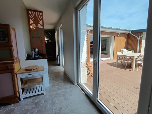 A corridor with a wooden table and chairs, leading to an outdoor patio area with a table and chairs.