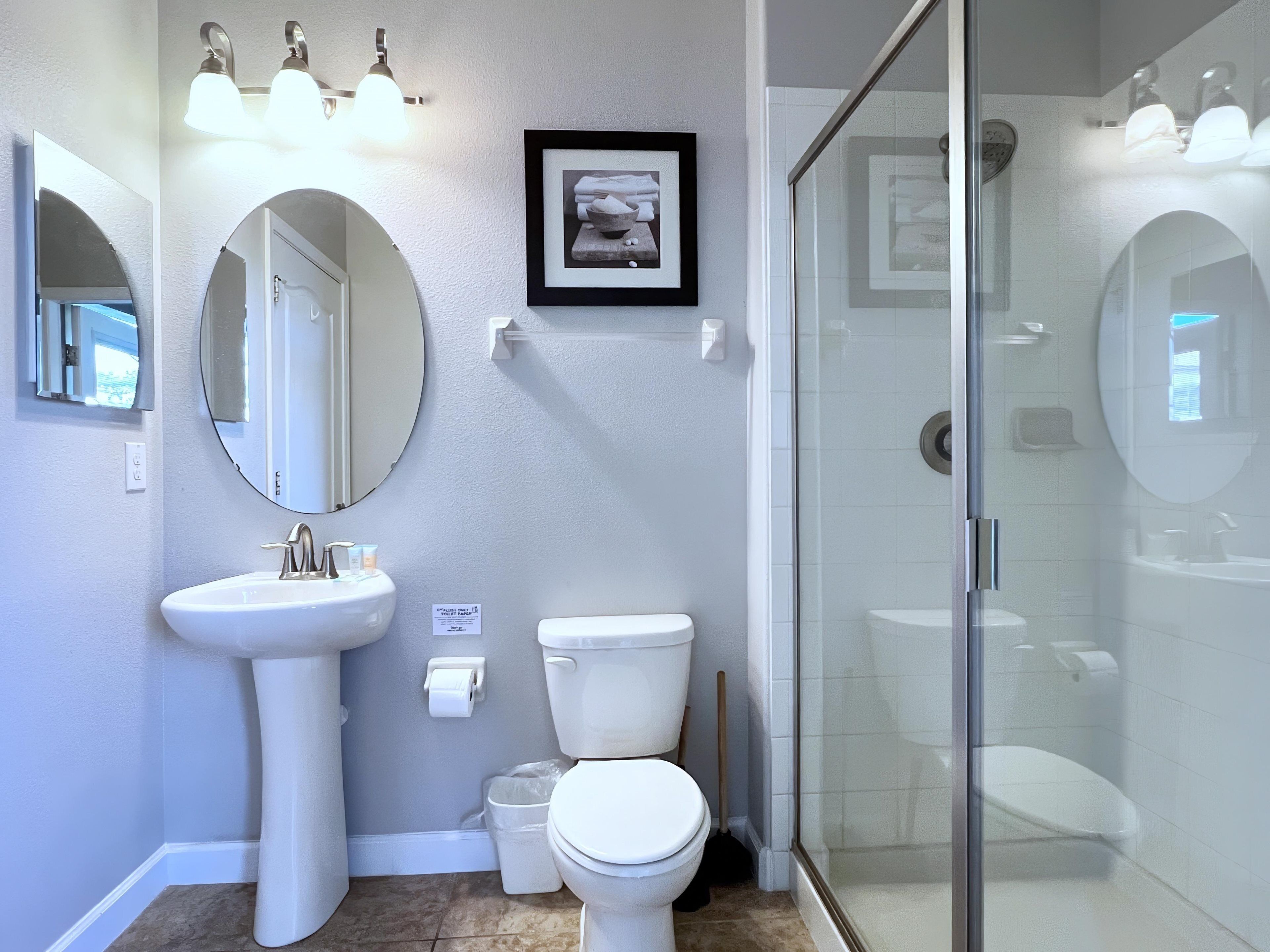 A bathroom with a pedestal sink, a toilet, a framed picture on the wall, and a glass shower enclosure.