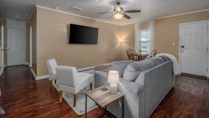 Ein Wohnzimmer mit grauem Sofa, einem weißen Sessel, einem kleinen Tisch mit Lampe, einem Deckenventilator und einem flachbildfernseher an der Wand.