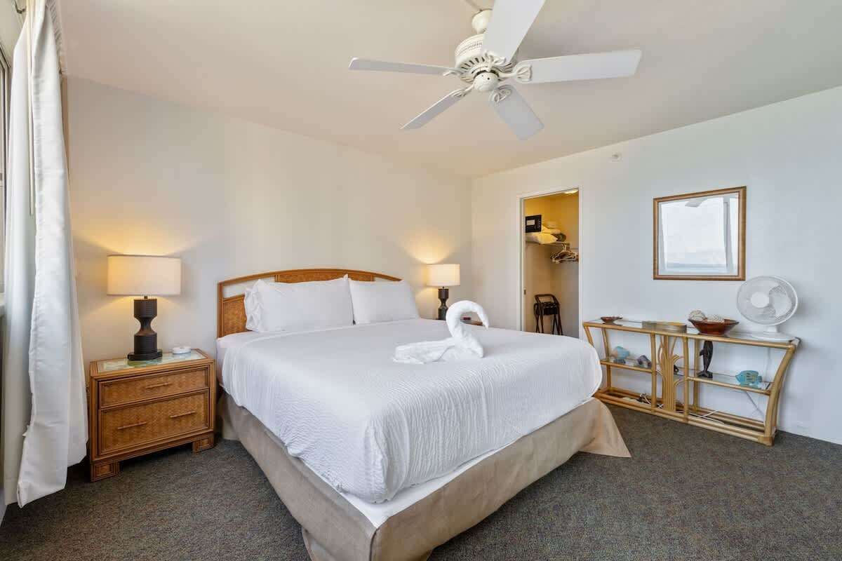 A hotel room with a bed, bedside lamps, a ceiling fan, a bedside table, a nightstand, a mirror, and a console table with a fan.