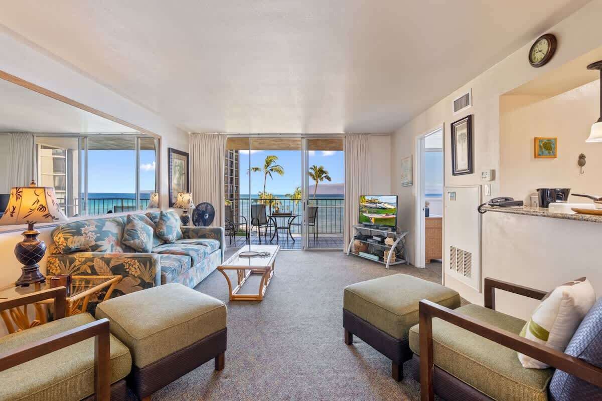 A living room with a sofa, ottomans, a coffee table, and a TV. There is a balcony with a view of palm trees and the ocean.