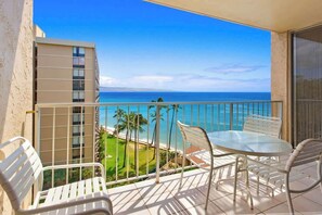 A balcony with a glass table and chairs, overlooking a beach and ocean.