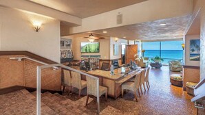 A spacious room with a long table, chairs, a desk, and a computer. There is a staircase with a railing, a large window with a view of the ocean, and a ceiling fan.