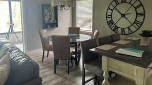 Un coin repas comprenant une table ronde, des chaises, un lustre, une grande horloge murale et une fenêtre avec des stores.
