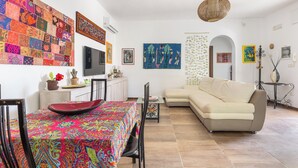 Una sala de estar con un colcha colorida, un sofá color beige, una mesa de comedor con sillas y un televisor.