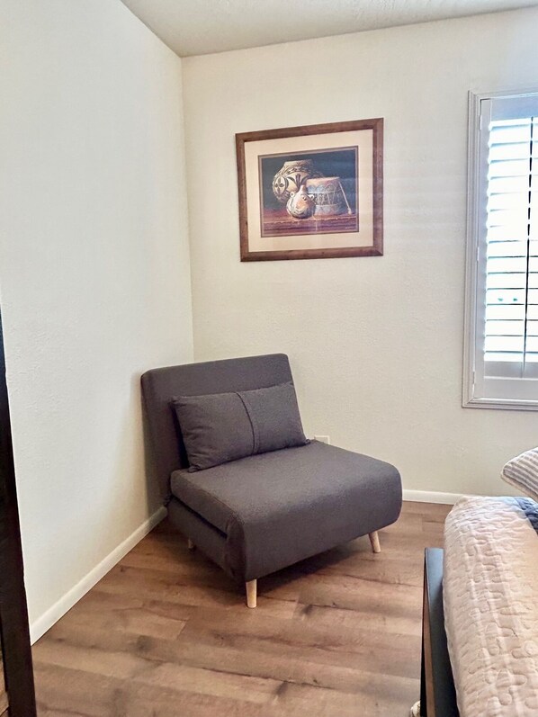 Um pequeno sofá de cor escura num canto do quarto, com um quadro na parede e uma janela com persianas.
