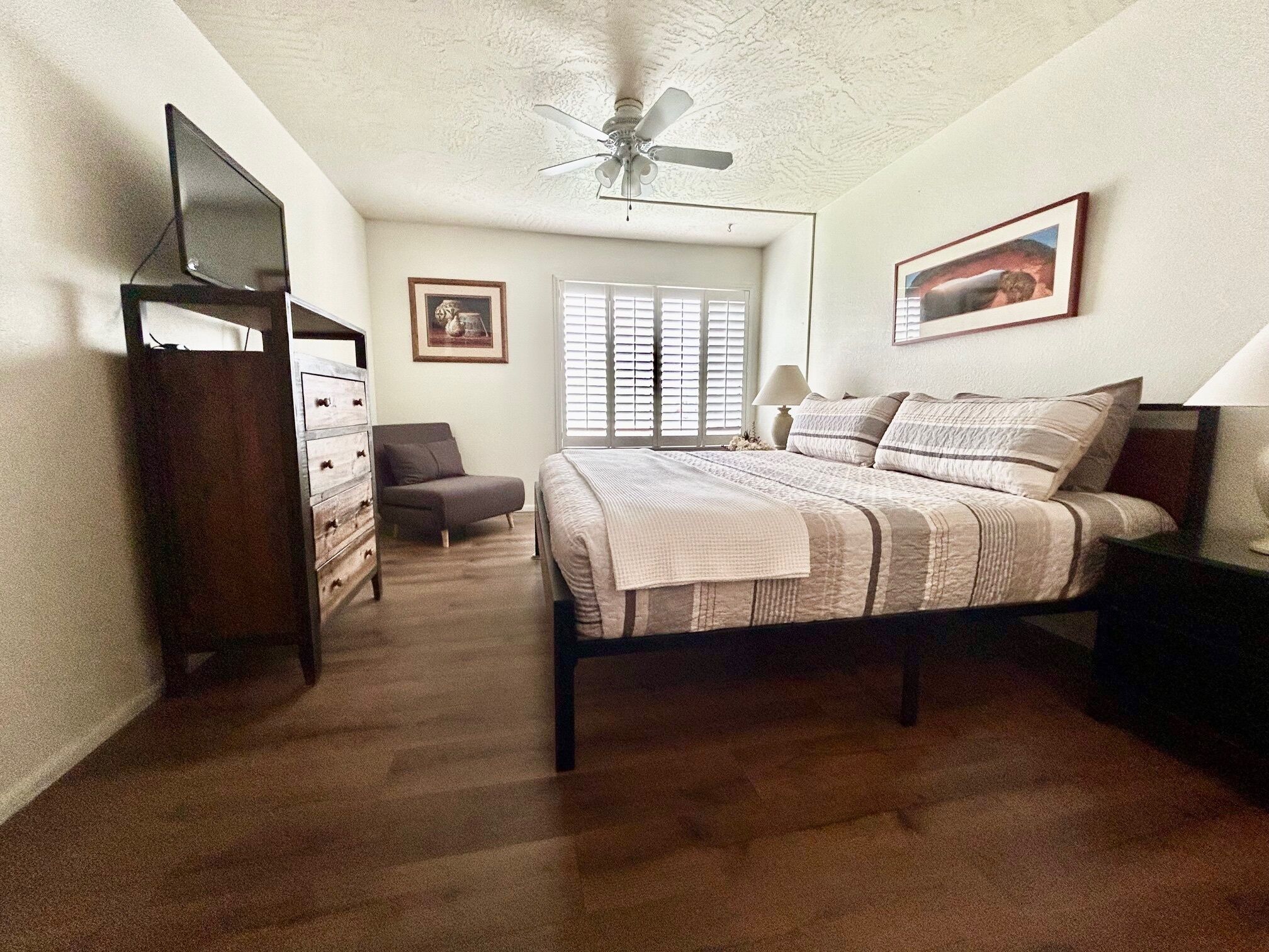 Une chambre à coucher comprenant un lit, une commode, une chaise, un ventilateur de plafond, une fenêtre avec des stores et un tableau encadré accroché au mur.