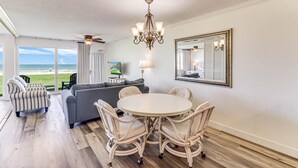 A spacious room with a large window overlooking the beach, a round dining table with chairs, a grey sofa, and a ceiling fan.