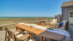 A wooden deck with a table and chairs, overlooking a beach and a building with a window. - Stunning Ocean View! Beachfront Nantucket Retreat (Nantucket)