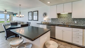A modern kitchen with white cabinets, a black countertop, and a tiled backsplash. There is a dining area with wicker chairs and a table. - Gulfstream Condos Unit 112 | Ground Floor | Gulf Front Views (Corpus Christi)