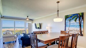 Zona pranzo con tavolo e sedie in legno, televisore a parete e un quadro che raffigura una spiaggia.