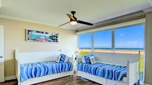 Una camera da letto con due letti bianchi, un ventilatore a soffitto, una grande finestra vista spiaggia e un quadro appeso al muro.