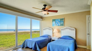 A bedroom with two beds, a ceiling fan, a nightstand with a lamp, and a view of the beach. - Gulfstream Condos Unit 117 | Pool & Hot Tub | Beachfront Patio (Corpus Christi)