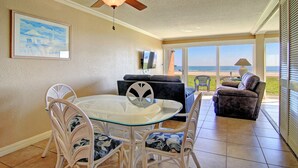 A dining area with a glass table and white chairs, a ceiling fan, a framed picture on the wall, and a view of the ocean through large windows. - Gulfstream Condos Unit 117 | Pool & Hot Tub | Beachfront Patio (Corpus Christi)