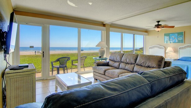 A living room with a large window overlooking the beach, a ceiling fan, and a view of the ocean. - Gulfstream Condos Unit 117 | Pool & Hot Tub | Beachfront Patio (Corpus Christi)