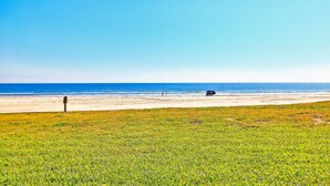 A beach with a clear sky, a sandy shore, and a grassy area in the foreground. - Gulfstream Condos Unit 117 | Pool & Hot Tub | Beachfront Patio (Corpus Christi)