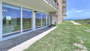 A modern building with large glass windows, a concrete pathway, and a grassy area leading to a beach. - Gulfstream Condos Unit 119 | Direct Beach Access from Gulf-View Patio | Pool (Corpus Christi)