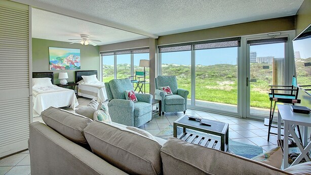 Amplia sala de estar con un sofá, dos sillones, una mesa de centro y una televisión. Amplias ventanas ofrecen vistas al exterior.