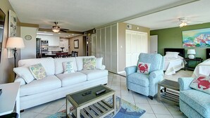 Una sala de estar con un sofá blanco, sillones y una mesa de centro. Se ve un comedor al fondo, así como una cocina.