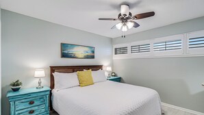 Une chambre à coucher comprenant un lit, deux tables de chevet, un ventilateur de plafond, un tableau et une fenêtre avec des stores.