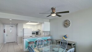 A modern kitchen with white cabinets, a central island, and a dining area with a glass table and wicker chairs. - Gulfstream Condos Unit 205 | Corner Unit with Beachfront Balcony (Corpus Christi)