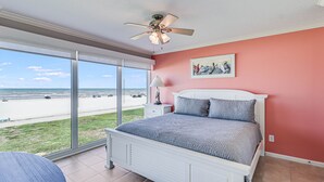 Een slaapkamer met een groot raam met uitzicht op een strand, een plafondventilator en een bed met een wit hoofdeinde.