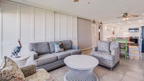 Een ruime woonkamer met een grijze bank, gestreept fauteuils, een ronde salontafel en een plafondventilator.