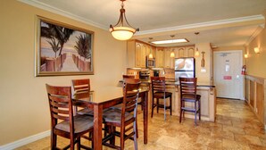 A dining area with a wooden table and chairs, a kitchen with wooden cabinets and appliances, and a painting of a beach scene on the wall. - Gulfstream Condos Unit 208 | Pool & Hot Tub | Private Balcony, Panoramic View (Corpus Christi)