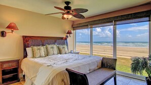 A bedroom with a large bed, a ceiling fan, a nightstand, a lamp, and a view of the beach. - Gulfstream Condos Unit 208 | Pool & Hot Tub | Private Balcony, Panoramic View (Corpus Christi)