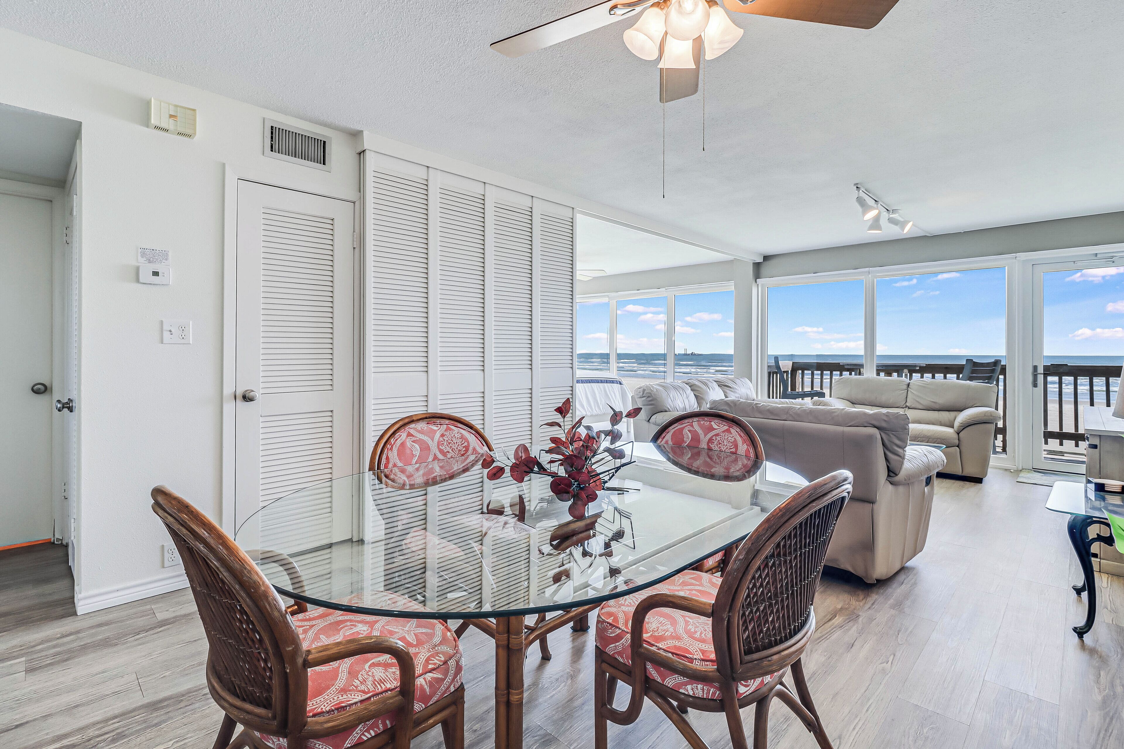 A modern living room with a glass dining table, wicker chairs, a beige sofa, and a view of the ocean.