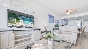 Un salon moderne doté d’un grand téléviseur à écran plat, d’une table basse en verre, d’un canapé blanc et d’un ventilateur de plafond.