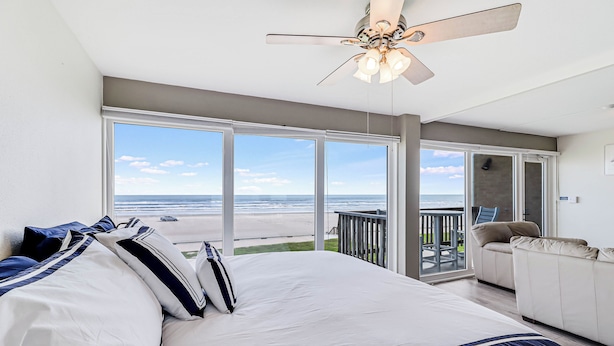 Une chambre avec un grand lit, un ventilateur de plafond et une vue sur la plage, visible à travers de grandes fenêtres.