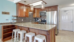 A kitchen with wooden cabinets, a large island with stools, stainless steel appliances, and a tiled backsplash. - Gulfstream Condos Unit 212 - Beach Front View & Balcony (Corpus Christi)