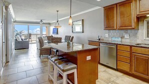 Una cocina amplia con una isla grande, armarios de madera y lavavajillas. Hay un comedor con mobiliario de mimbre y vistas al exterior.