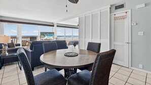 A dining area with a round table and chairs, large windows with a view of the beach, and a ceiling fan. - Gulfstream Condos Unit 215 | Beachfront Balcony | Pool & Hot Tub (Corpus Christi)