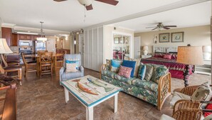 A spacious living area with a dining table, chairs, a sofa, and a coffee table with a fish design. There are ceiling fans, a kitchen in the background, and a bed with a red bedspread. - Gulfstream Condos Unit 216 | Direct Gulf Views | Pool, Hot Tub (Corpus Christi)
