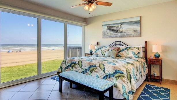 A bedroom with a large window, a bed with a tropical print bedspread, a bench, and a ceiling fan. - Gulfstream Condos Unit 217 | Gulf-Front View with Beach Balcony & Pool (Corpus Christi)