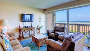 A living room with a sofa, armchairs, a coffee table, and a TV on the wall. There is a large window with a view of the beach. - Gulfstream Condos Unit 217 | Gulf-Front View with Beach Balcony & Pool (Corpus Christi)