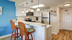 Una cocina con armarios blancos, un refrigerador de acero inoxidable, un microondas y una cafetera. Hay dos taburetes de mimbre con cojines coloridos.