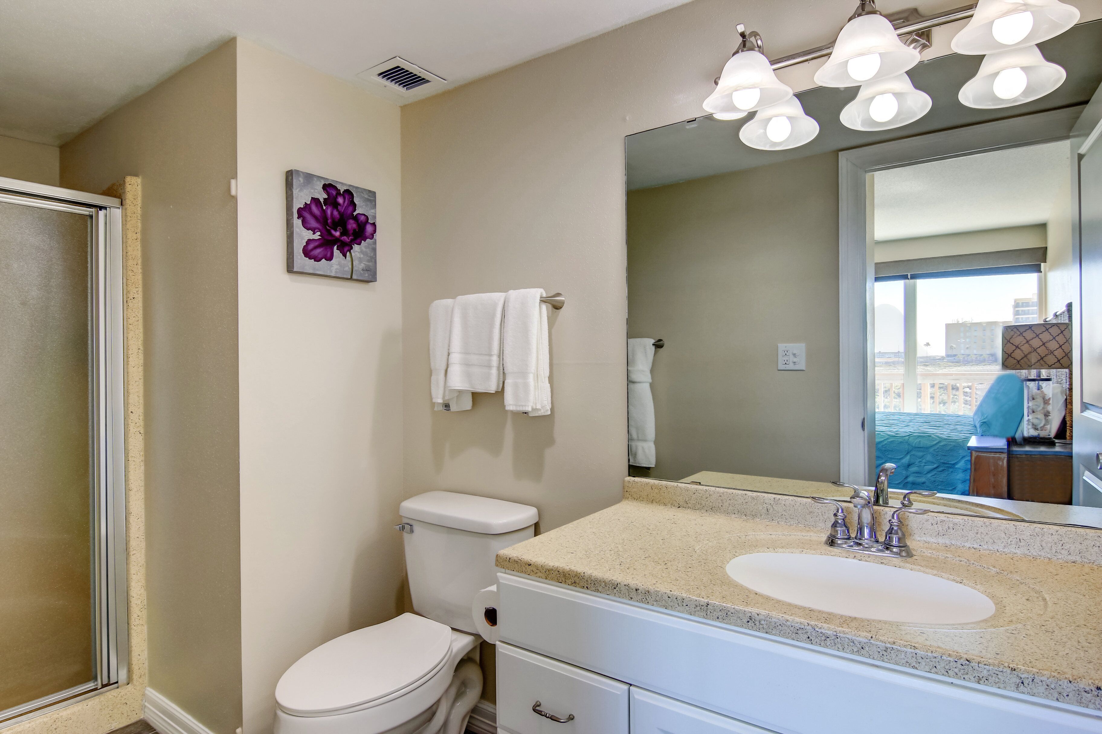 A bathroom with a toilet, sink, and shower. There is a mirror above the sink and a painting on the wall.