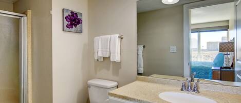 A bathroom with a toilet, sink, and shower. There is a mirror above the sink and a painting on the wall.