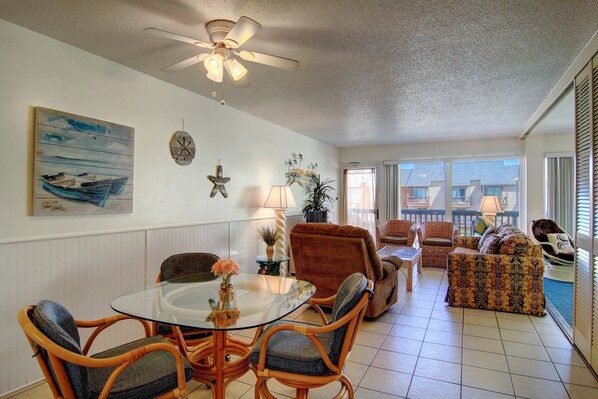 A spacious living area with a glass-top dining table, wicker chairs, a sofa, and a ceiling fan. - Gulfstream Condos Unit 301 | Extended Private Balcony | Pool, Hot Tub & BBQ (Corpus Christi)