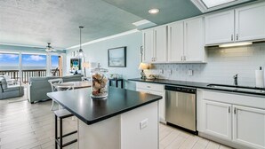 A modern kitchen with white cabinets, a black countertop, and a dishwasher. There is a dining area with a view of the ocean. - Gulfstream Condos Unit 306 | Gulf Front Views & Balcony | Pool, Hot Tub (Corpus Christi)