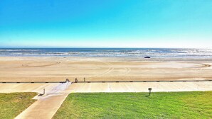 A wide beach with a clear sky, a sandy path leading to the water, and a grassy area in the foreground. - Gulfstream Condos Unit 312 | Balcony with Beach Front Views, Pool & Hot Tub (Corpus Christi)