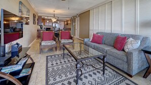 Een ruime woonkamer met een grijze bank, twee fauteuils, een glazen salontafel en een tapijt met een patroon. Een plafondventilator en hanglampen zijn zichtbaar.