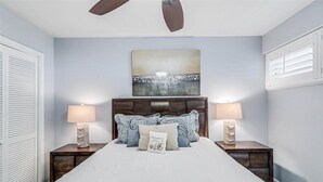 Une chambre à coucher avec un lit, deux tables de chevet avec des lampes, un ventilateur de plafond, un tableau accroché au mur et une fenêtre avec des stores.