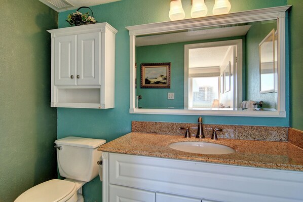 A bathroom with a white toilet, a granite countertop sink, and a large mirror above the sink. - Gulfstream Condos Unit 320 | Beach-View Balcony | Heated Pool, Hot Tub (Corpus Christi)